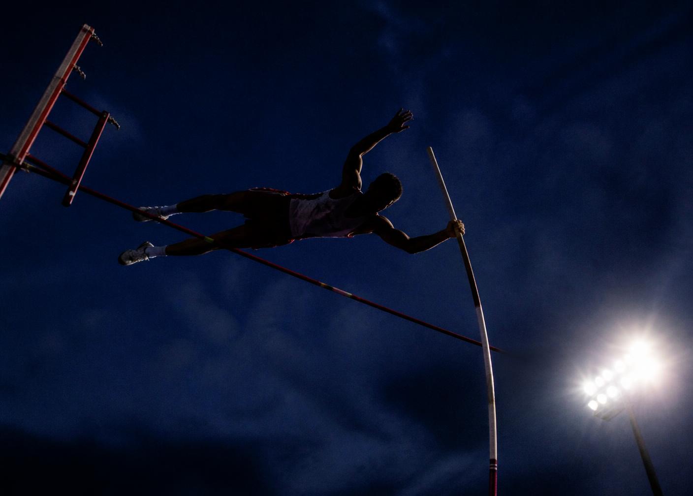 Illustrated night scene of a vaulter clearing the bar under stadium lights